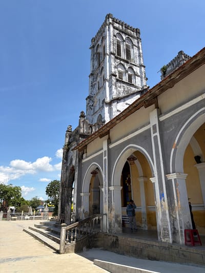 A wide-angle, eye-level outdoor shot captures the exterior of a weathered church in Hội Tín, Vietnam. The church features a tall, stone bell tower with arched windows and a crenellated top, showing signs of age and neglect with peeling paint and cracks. To the right of the tower, a covered walkway with arched openings leads into the building. The walkway is supported by columns and features a yellow-painted interior. A few people are visible: one man in a denim jacket stands inside the arched walkway, appearing to be on his phone, while another person in darker clothing stands further inside. A red plastic stool is placed outside the walkway. In the foreground, a wide, concrete staircase with a stone balustrade leads up to the church entrance. The ground in front of the church is paved concrete, with some bare ground and greenery visible further in the distance. A string of small lights hangs across the scene under a bright blue sky with scattered white clouds. The overall impression is of an old, historical religious structure in a tropical climate.