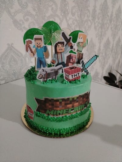The scene depicts a Minecraft-themed celebratory cake situated on a light-colored surface within an indoor environment in the city of Eshmataul, Uzbekistan. The single-tier round cake is frosted in a vibrant green, with green piped frosting resembling grass along its base and top edges. The side of the cake displays a rectangular, pixelated image depicting a dirt block texture with the word "MINECRAFT" centrally placed. The top of the cake features various paper cutouts, including Minecraft characters Steve (holding a pickaxe), Jesse (holding a sword), and Alex, alongside a pig, a wolf, and a diamond sword. Two red-and-white "TNT" block cutouts are also positioned on the cake's surface, with an additional smaller "TNT" cutout on the lower left side. Several translucent green lollipops, shaped like circular foliage, are interspersed among the character toppers. The cake rests on a gold-colored circular cake board. The background consists of a light-toned wall covered with subtly reflective, patterned wallpaper featuring a repeating ornamental design. A dark-leafed plant is partially visible in the far right background, and a faint shadow extends from the cake to the right across the surface it rests upon.