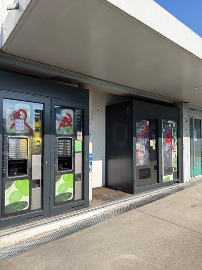The image depicts a row of outdoor vending machines situated under a concrete overhang in the city of Saint-Priest, France. Two prominent dark gray vending machines on the left feature green decorative panels, glass fronts, and visible selection interfaces with coin/card slots. Both of these machines display a red spray-painted hammer and sickle symbol on their glass fronts. The leftmost machine shows some yellow discoloration near its upper display. To the right, a larger, darker gray vending unit, possibly comprising multiple machines, also features clear glass fronts revealing various products inside. This unit is likewise marked with red spray-painted hammer and sickle symbols and additional indecipherable graffiti, with a large black rectangle obscuring part of its left panel. The machines are positioned against a light-colored wall. A robust concrete overhang provides shelter above the machines, supported by a dark beam. The ground in the foreground consists of light-colored pavement with a long, perforated metal drainage grate running horizontally. The scene is illuminated by daylight, with a clear blue sky visible in the upper right quadrant. No people are present.