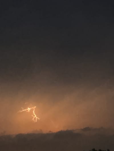 A lightning bolt illuminates the sky above Putten, Netherlands. The lightning is bright yellow and forks out in multiple directions. The sky is dark and cloudy, with a hint of orange light emanating from below the storm clouds. The bottom of the image shows a dark silhouette of trees.