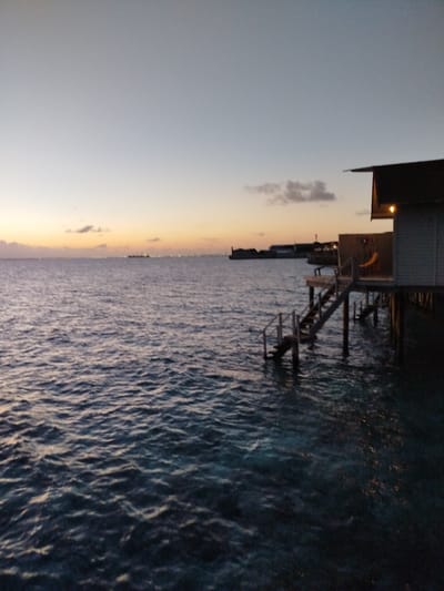 This is a full-frame, horizontal, medium-shot, outdoor, eye-level, natural-light photograph taken at dusk in Gulhi Falhu, Maldives. In the foreground, dark blue, choppy ocean water fills the bottom half of the frame. A wooden staircase with railing descends from a building on stilts, on the right side of the frame, into the ocean. The building has a brown roof and white siding, and a light is visible inside. In the middle ground, a distant shoreline with buildings and lights is visible across the ocean. The sky is a gradient of pale blue and orange, with a few clouds on the left. The overall mood is serene and tranquil.