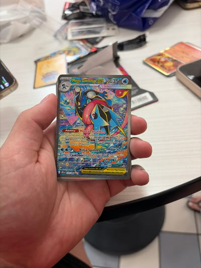 A person's hand holds a "Mega Greninja-EX" Pokemon card. The card is silver-bordered and features a colorful illustration of the Pokemon. The card's text is visible, including its HP of 350, abilities "Mortal Shuriken" and "Ninja Spinner," and its attack damage of 120+. The background of the image is blurred, showing what appears to be a table with other Pokemon cards and packaging. The scene is indoors, possibly in a room or a store. The location is unknown, as no specific landmarks or identifying features are visible.
