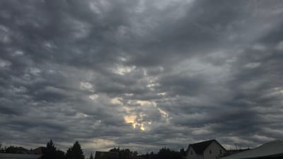 The image captures a dramatic, overcast sky above a residential area in Strokino, Russia. Heavy, dark grey clouds dominate the frame, with a patch of bright, golden light breaking through the clouds in the upper center, suggesting either a break in the storm or a strong sunset/sunrise. Below the sky, the rooftops of several houses are visible, interspersed with dark green evergreen trees and other foliage. One house in the lower right quadrant has a prominent dark roof and light-colored siding. The overall atmosphere is somber and foreboding, indicative of approaching or passing inclement weather. There are no people or animals visible in the scene.