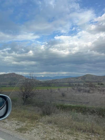 The image displays a wide landscape view, likely captured from a moving vehicle, evidenced by a silver side mirror visible in the lower-left quadrant. The scene is located near Ribnik, Bulgaria. The sky occupies approximately two-fifths of the frame, presenting a mix of blue patches and extensive white to dark grey cumulus and stratocumulus clouds. Four parallel power lines traverse horizontally across the upper-middle portion of the sky.
In the background, a range of snow-capped mountains is visible in the distance, positioned above a series of rolling, largely barren hills. The midground consists of an expanse of dry, dormant vegetation, featuring numerous leafless deciduous trees and shrubs, indicative of late winter or early spring. Interspersed within this dry landscape are distinct, vibrant green agricultural fields. The immediate foreground shows dry, patchy ground cover, consistent with a roadside verge. No human figures or direct interactions are depicted within the scene.