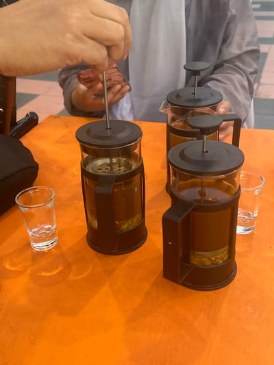 The image depicts a group of people preparing and serving tea using French presses on an orange table, likely in a cafe or outdoor seating area in Bantaeng, Indonesia. In the foreground, three glass French presses are visible, two of which are partially filled with a golden-brown liquid and visible tea leaves or herbs at the bottom. The plungers of the French presses are in various positions, with one being actively pressed down by a hand, suggesting the brewing process is underway. Two small, clear shot glasses are also present on the table, one to the left of the French presses and another to the right, presumably for serving the tea. In the background, the arm of a person holding a smartphone is visible, and another person is wearing a grey garment. The table surface has a textured, wood-grain appearance. The lighting suggests it is daytime.