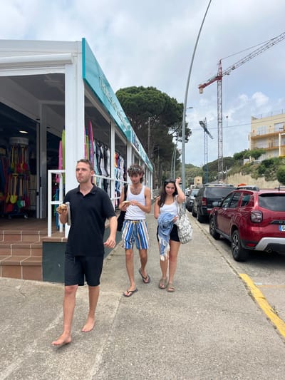 The scene is a street in Tamariu, Spain, located at coordinates 41.919, 3.206. Three people are walking down the street. Two men are walking side-by-side in the middle of the sidewalk. The man on the left is barefoot and wearing black shorts and a black t-shirt. He is holding a small, brown paper bag in his left hand. The man on the right is wearing white tank top, blue and white striped shorts, and sandals. He is holding a piece of food in his right hand. A woman is walking behind the men and slightly to the right. She is wearing a white tank top, a blue and white striped skirt, sandals, and has a blue and white striped beach towel slung over her left shoulder. The woman is carrying a white and gray tote bag in her right hand. There is a building on the left side of the scene, with a large, blue and white sign that says "THEMES SHOP." The building has a large window with a variety of items for sale inside. There is a large, white van parked on the right side of the scene. The road is paved with gray concrete. There is a large construction crane in the background. The sky is blue with a few clouds. The weather is sunny and warm.