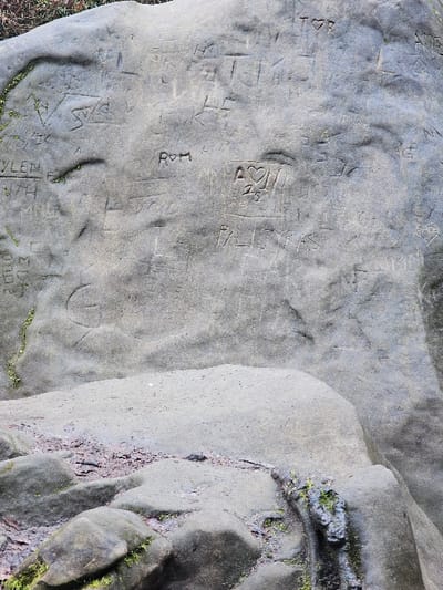 The image displays a large, weathered sandstone rock face situated in Waldbillig, Luxembourg. The prominent greyish-brown rock surface is extensively covered with numerous carved inscriptions, including initials, names, and symbols, indicating deliberate human alteration. Notable carvings include "ROM," "PALOVAS," "T♡R," "VSA," and an "A♡T" symbol enclosed within a square, with the number "25" positioned below it. Patches of green moss adhere to various crevices and lower, damp sections of the rock. The base of the rock features accumulated dark, reddish-brown soil and organic debris within its depressions. No individuals are present in the scene, and no active interactions are depicted.