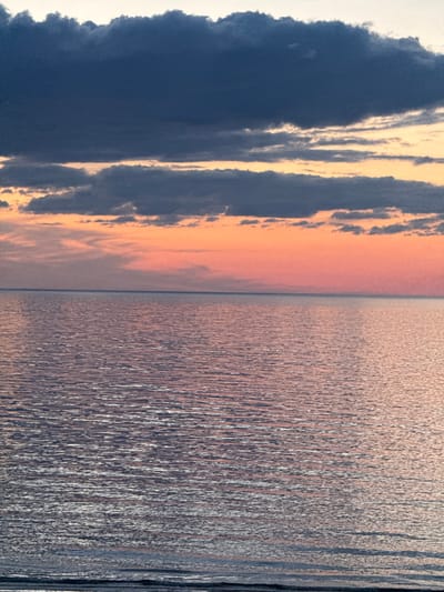 The image shows a body of water meeting the sky at the horizon. On the left of the image, there are dark clouds, with lighter orange hues peaking through them. The water surface on the right reflects the colors of the sky. There are no people or objects visible. The location is presumed to be Kalngale, Latvia, based on the instructions.