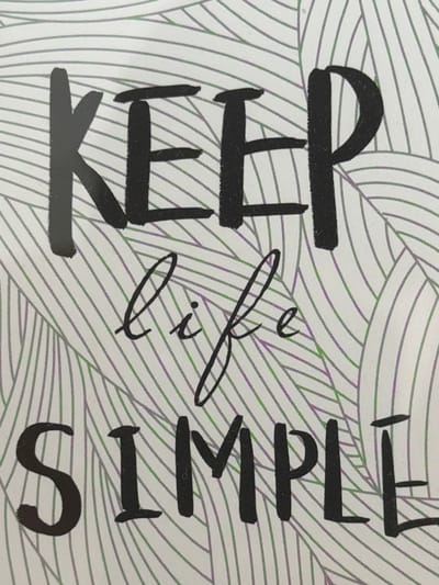 The image is a close-up photograph of text on a patterned background. The text reads "KEEP life SIMPLE". "KEEP" and "SIMPLE" are in large, bold, black, sans-serif capital letters. "life" is written in smaller, cursive, black script. The background consists of a white surface with a pattern of thin, curved, parallel lines in green and purple, resembling stylized leaves or waves. The image is cropped tightly and does not show any environment or context beyond the text and its immediate background. There are no people, actions, or interactions depicted. The image does not provide any information about the city of Spinea, Italy.