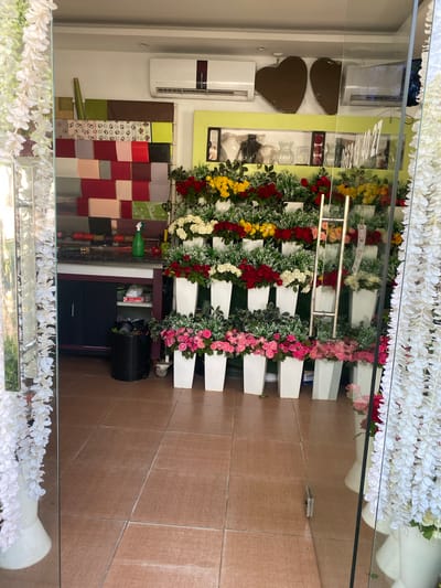 The image captures a retail display, likely a florist or gift shop, in Sidi Moussa, Algeria. The scene is dominated by an impressive arrangement of artificial flowers, predominantly in white, red, yellow, and pink, cascading down a tiered white structure. These floral displays are the main subject, creating a vibrant and festive atmosphere.

Behind the flowers, several objects are visible, offering clues about the shop's purpose. These include what appear to be decorative heart-shaped items, possibly gifts or confectionery. Further back, a white air conditioning unit is mounted on the wall, and a display of colorful wrapped fabric squares is arranged on a rack, suggesting the shop might also sell textiles or gift-wrapping supplies. A black plastic bin is also noticeable.

The reflection in the glass storefront indicates the presence of an outdoor space, with the floor tiles suggesting an indoor tiled area. The lighting appears bright, hinting at daytime. There is no visible text in the image that can be deciphered. The overall impression is of a well-decorated shop prepared to showcase its products.