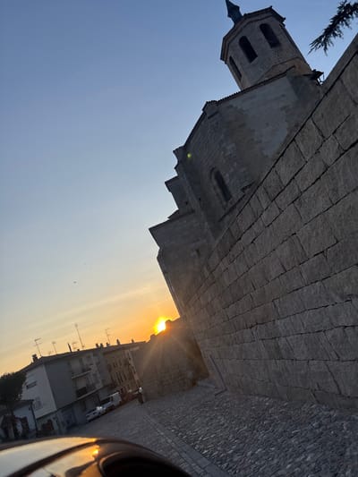 The image depicts a sunset scene in Ávila, Spain, specifically featuring the Basilica de San Vicente. The perspective is angled upwards and to the right, highlighting the robust stone architecture of the basilica.
The upper right portion of the frame is dominated by the Basilica de San Vicente, a large, historic stone structure with walls composed of precisely cut grey blocks. A prominent bell tower, featuring arched openings and a pointed spire, rises from the main building. A dark green pine branch extends into the upper right corner, partially obscuring the tower.
The sky, transitioning from a light blue at the zenith to a vibrant orange and yellow on the horizon, indicates the setting sun, which is visible as a brilliant, radiating orb near the center-left horizon, partially obscured by the basilica's lower wall and a distant building, causing a significant lens flare. Several small, indistinct birds are visible in the mid-left sky.
A dark grey cobblestone street extends from the bottom of the frame, curving slightly towards the left. On the left side of the street, a row of multi-story buildings, consistent with residential or commercial structures, displays windows, balconies, and multiple television antennas on their rooftops.
Two dark, silhouetted human figures are discernible on the street in the mid-ground, near parked vehicles. A white commercial van and at least one other vehicle are parked or moving on the street between the modern buildings and the basilica wall. The immediate foreground, in the bottom left, shows a dark, reflective surface, likely a vehicle's dashboard or hood, reflecting the golden light of the sunset.