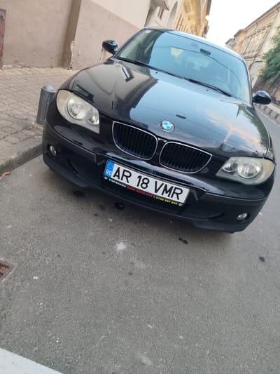 A black BMW is parked on a street in Arad, Romania. The car is facing forward, and its license plate reads "RO AR 18 VMR". The street is paved with asphalt, and there is a sidewalk made of cobblestones to the left of the car. The buildings in the background appear to be old and ornate. The sky is overcast, suggesting it is either early morning or late afternoon. The car is the primary subject of the image, and its details are well-captured. There are no people visible in the image. The overall scene is typical of a city street in Romania.