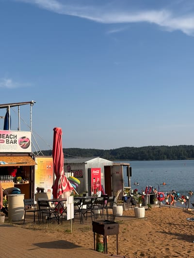 This is a vertical, eye-level shot of a beach on a sunny day. The foreground is dominated by a sandy beach, with a wooden walkway and a beach bar. The bar has a red umbrella, tables, chairs, and several signs, including one advertising "BEACH BAR DUBROVO BROVO" and another with "Coca-Cola" branding and a menu. There are also some decorative elements like white planters and a shopping cart. Further back, a line of people are relaxing on the shore and in the water, which is calm and reflects the clear blue sky. A dense line of trees borders the far side of the water. The overall atmosphere is relaxed and summery, suggesting a popular spot for recreation and leisure. The bright sunlight and clear sky indicate it's likely midday or early afternoon.