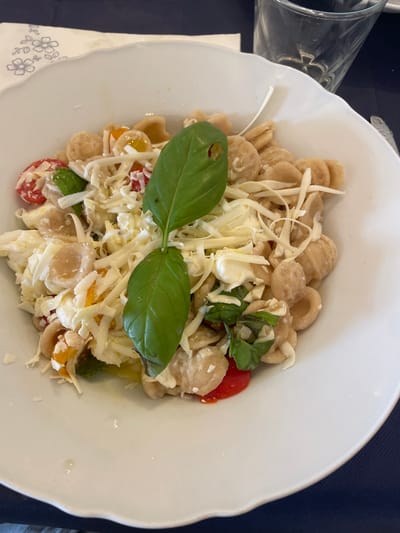 A white scallop-edged bowl, located in Ceglie Messapica, Italy, contains a pasta dish. The dish is composed of light brown orecchiette pasta, topped with shredded white cheese, small cubes of fresh mozzarella, halved red and yellow cherry tomatoes, and fresh green basil leaves. One prominent basil leaf in the center displays a small, irregular perforation. A subtle sheen consistent with olive oil is visible at the bottom of the bowl. The bowl rests on a dark blue tablecloth. A white paper napkin with a light blue floral pattern is partially visible to the upper left of the bowl, and a clear glass of water is partially visible to the upper right. A silver utensil handle is barely visible on the far right edge. No individuals or direct actions are depicted.
