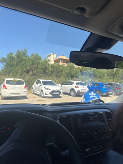 The image depicts a scene viewed from inside a parked vehicle in Georgioupoli, Greece, looking outward through the windshield on a clear, sunny day. The sky is cloudless and a vivid blue. In the mid-ground, a row of five cars is parked on a light-brown, unpaved surface, likely a dirt or gravel lot, with dense green trees and bushes forming a backdrop. Behind the foliage, light-colored buildings with flat roofs are visible.
From left to right, the parked vehicles are:
1. A white Hyundai i20 hatchback, viewed from the rear, displaying the Greek license plate "XMK-3743".
2. A white Toyota Yaris hatchback, positioned diagonally, showing its front-left side, with the Greek license plate "KMO-1339".
3. A white Citroën C3 Aircross, visible from its rear-left quarter.
4. A blue Peugeot 208 hatchback, viewed from the rear, with a partially visible Greek license plate beginning with "YAB-".
5. A dark grey or black SUV, seen from its rear-right side, parked further back.
The interior of the observer's vehicle features a dark-colored dashboard and a partial view of the steering wheel on the left. A blue Adidas cap with a white trefoil logo is placed on the dashboard directly beneath the rearview mirror, which itself reflects part of the interior and the exterior scene. The infotainment system in the center console displays "TEL 210" and "92.90 MHz" on the top line, with "20.5" (likely temperature) on the bottom right. A fragment of dark hair belonging to a passenger is visible on the far right edge of the frame. The windshield exhibits a dotted frit band along its top edge and around the rearview mirror housing.