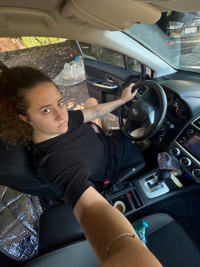 The image shows a young woman inside a car, with her right hand on the steering wheel. She is wearing a black t-shirt and appears to be looking towards the camera. The car's interior is visible, including the dashboard, steering wheel, gear shifter, and cup holder. Outside the car, trees and foliage are visible, suggesting a natural environment. The license plate of a vehicle behind her is partially visible and reads "TX9663AH". It's not possible to confirm the location as Durankulak, Bulgaria from the image alone.