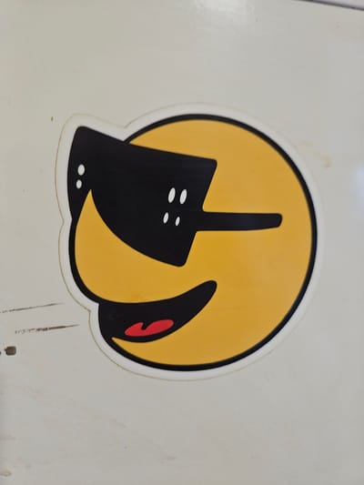 The image shows a sticker of a yellow smiley face wearing black sunglasses and a red mouth, applied to a white, textured surface. The smiley face has two white dots for eyes within the black sunglasses. The sticker is slightly raised from the surface. There are some faint markings and smudges on the white surface surrounding the sticker. The scene appears to be indoors or under a covered area, possibly a wall or a piece of equipment, given the uniform lighting and lack of distinct environmental features. The image has not been geo-tagged with the city of Arnhem, Netherlands.