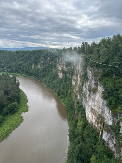This image shows a sweeping view of a lush green forest bordering a wide, brown river. A sheer, rocky cliff face, partially covered in vegetation, rises from the forest on the left side of the frame. A suspension bridge, made of cables and wooden planks, is visible spanning across the cliff and forest. A plume of mist or fog rises from the cliff face near the bridge, suggesting a waterfall or humid conditions. The sky is overcast with heavy, grey clouds, indicating a cloudy or possibly rainy day. The light is diffused and the overall atmosphere appears serene and natural. The photo is taken from a high vantage point, looking down and across the river valley.