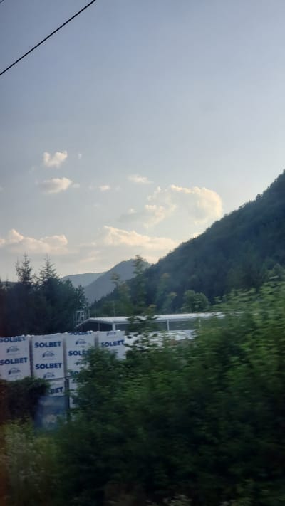 The image depicts a landscape scene in the vicinity of Ľubochňa, Slovakia. In the foreground, stacks of white building materials, possibly insulation boards, are visible. These materials are marked with the brand name "SOLBET". Adjacent to these stacks are dense green trees and foliage. In the midground, there is a white building structure with a roof, partially obscured by trees. The background features rolling hills and mountains covered in dark green forests, under a pale blue sky with scattered white clouds. A thin black line, likely a power line, stretches across the upper left portion of the image. The overall impression is one of a rural or industrial area bordering a natural mountainous environment.