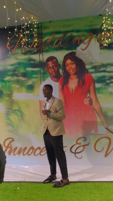 A man wearing a tan blazer, white shirt, and dark pants stands in front of a wedding backdrop. The backdrop features a couple, a woman in a red dress and a man in a white shirt, against a blurred green background with the words "Wedding" and "Innocence & V." in cursive lettering. String lights are draped on the side of the backdrop. The man in the blazer is positioned in front of the couple's image on the backdrop.