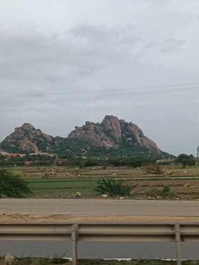 The image shows a panoramic view of a rocky mountain range with lush green vegetation covering its slopes. The sky is overcast with a layer of clouds. In the foreground, there is a road with a metal barrier. Beyond the road, there are agricultural fields and scattered trees. The overall scene appears to be a rural landscape with natural beauty. The image is a photograph taken outdoors, capturing a wide shot of the scenery. The location is Addakal, India.