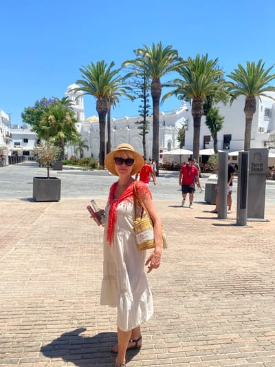 A light-skinned woman with blonde hair and sunglasses, wearing a white tiered dress, a straw hat, and a red scarf, stands in a brick-paved plaza under a bright blue sky. She is holding a bottle of water and a phone. In the background, palm trees and white buildings with arched windows and a dome are visible. Several people are walking in the plaza, and outdoor seating areas with umbrellas are set up in front of some buildings. A sign reads "Torre de Guzman." The scene appears to be daytime in a warm climate, likely the town of Conil De La Frontera, Spain, given the architecture and signage.