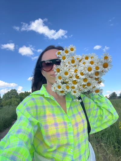 A young woman with dark hair and sunglasses is holding a large bouquet of daisies in front of her face, obscuring most of her features. She is wearing a bright green and purple plaid shirt and is standing outdoors in a field with green grass and trees in the background. The sky is blue with scattered white clouds. The image appears to be a selfie taken on a sunny day. This scene is located in the city of Pigozino, Russia.