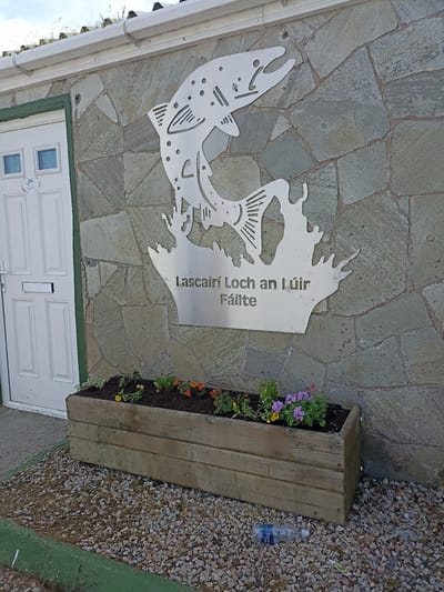 The image shows the exterior of a building with a stone facade in Loughanure, Ireland. A metal sculpture of a leaping fish adorns the wall, with text below it that reads "Lascairí Loch an Lúir Fáilte". In front of the building, there is a wooden planter box filled with soil and various small flowering plants, including purple, yellow, and orange varieties. A white door with two small windows is visible to the left of the sculpture. The ground in front of the planter is covered with small grey stones. A plastic water bottle and a piece of wire are on the ground near the planter. The building has a white gutter and a roof with green vegetation growing on it.