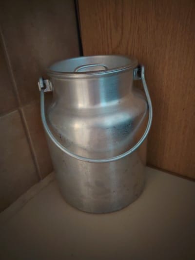 An aluminum milk can with a handle sits in a corner. The can is mostly silver and appears slightly tarnished in places. The background consists of a tiled wall on the left and a wooden surface on the right. The scene is lit from above and to the right, casting a shadow to the left.