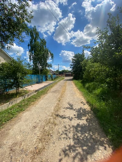 The image depicts a dirt road leading into a residential area in Akhtyrskiy, Russia. The road is lined with vegetation, including trees and grass, and appears to be well-traveled. On the left side of the road, there is a blue fence and a house. On the right side, there are more trees and foliage. In the background, there are houses and a gate. The sky is blue with scattered white clouds, and the sun is shining. The overall impression is one of a quiet, rural setting.