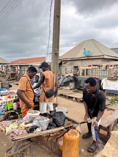 In Oko Amakom, Nigeria, under a cloudy sky, a market scene unfolds. Three men are engaged in commerce around a cart laden with various goods. The man on the left, wearing an orange polo shirt and orange shorts, is bent over the cart, examining items. A black crossbody bag hangs on his shoulder. To his right, a man in an orange sleeveless tunic and white shorts, wearing a black baseball cap, stands and appears to be interacting with the man in orange. He also carries a black crossbody bag. Behind them, another man in a blue and white striped shirt looks on.
The cart itself displays an assortment of products. Two large clear plastic bags filled with small, light-brown items are prominent, as are smaller bags containing similar contents. Various other small items, some wrapped in plastic, are also visible. To the right of the cart, a man in a black t-shirt and white shorts is actively engaged, holding a blue plastic bag and a white object. He wears a bracelet on his left wrist and dark sandals. A large, orange plastic container is situated near his feet.
The environment suggests a bustling outdoor market. Wooden structures and makeshift stalls are visible in the background. Pallets and tarps are used for display and storage. A concrete utility pole with wires runs vertically through the scene, and a satellite dish is mounted on a building's roof. The ground is dirt and debris, indicating a well-trodden market area.