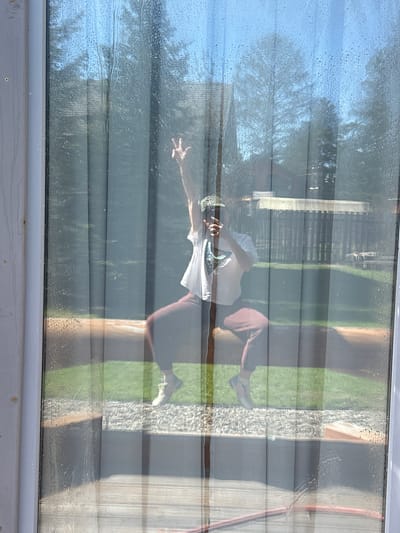 The image shows a person reflected in a rain-streaked window. The person is wearing a white t-shirt and reddish-brown pants, with one arm raised. The reflection also captures the outdoor environment, including grass, trees, and a building with siding.