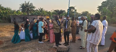 The image captures an outdoor gathering of approximately 20-30 individuals in Alayi, Nigeria, during daylight hours under a clear sky. The scene features a transition from dry, reddish-brown unpaved ground on the left to a paved road segment on the right, separated by a visible concrete drainage culvert.
On the left side of the frame, near the unpaved ground, a group consisting of women and young girls is present. Four adult females are attired in identical long, teal-colored formal gowns. One young girl also wears a similar teal dress, while another small girl with reddish-brown curly hair is dressed in a white formal gown. Behind this group, a single-story dilapidated building constructed of cinder blocks is visible, backed by dense tropical vegetation including banana or plantain trees and a tall palm tree.
In the center of the image, two men stand on the concrete culvert cover. Both are dressed in black trousers, white dress shirts, black ties, and distinctive long-sleeved jackets featuring a gold and black leopard-print or abstract pattern. The man on the right of this pair wears a black cap with white lettering, appearing to read "PDC." Both individuals are looking towards the right side of the frame, exhibiting slight smiles.
To the right, on the paved road, a larger group of people is assembled. Their clothing varies, encompassing traditional Nigerian attire and semi-formal wear. One man in a light-colored (cream/beige) traditional outfit with a black patterned scarf is observed looking down at a mobile phone. A woman, facing away from the camera, is visible in a white shirt with thin vertical reddish-brown stripes and matching trousers, carrying a black handbag. Partially obscured behind this group on the far right is a white van. A blue utility pole or street light standard is also present on the far right midground.