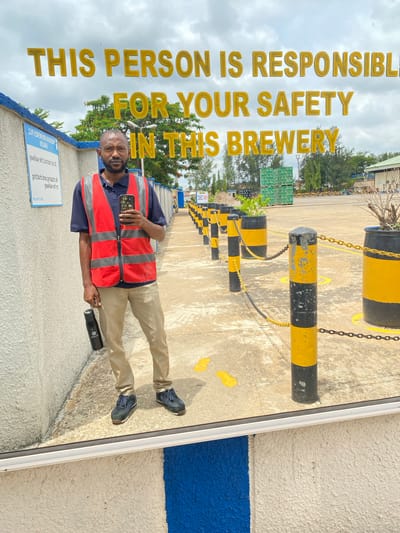 The image shows a man standing in what appears to be the grounds of a brewery in Kinkinau, Nigeria. He is wearing a navy blue polo shirt, tan pants, a red and gray reflective safety vest, and blue sneakers. He is holding a black water bottle in his right hand and has a smartphone clipped to his vest.
Behind the man, large yellow and gold lettering on a reflective surface reads: "THIS PERSON IS RESPONSIBLE FOR YOUR SAFETY IN THIS BREWERY". This suggests a focus on safety protocols within the facility. The background shows a paved area with yellow and black striped bollards and chains, common safety features in industrial settings. There are also stacks of green crates visible in the distance, likely containing the brewery's products. Trees and a cloudy sky are also part of the background, indicating it is daytime.
A small blue and white sign is visible on a wall to the left of the man, with some text on it. Another sign with text is partially visible in the far background. The man's expression is neutral, and the overall scene appears to be of a typical workday at an industrial facility.