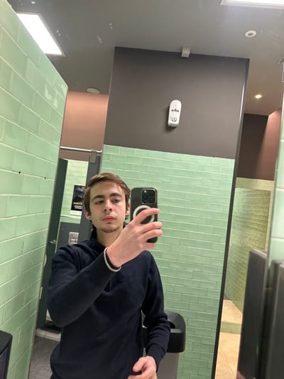 A young man takes a selfie in a bathroom mirror. The room features light green, rectangular tiles covering the lower walls. Above, the walls are dark grey, with a brown section visible in the upper corner. A white object, possibly a sensor, is mounted on the dark grey wall. The man wears a dark blue sweater and a gold bracelet. His phone, with a visible MagSafe ring, obscures part of his face.