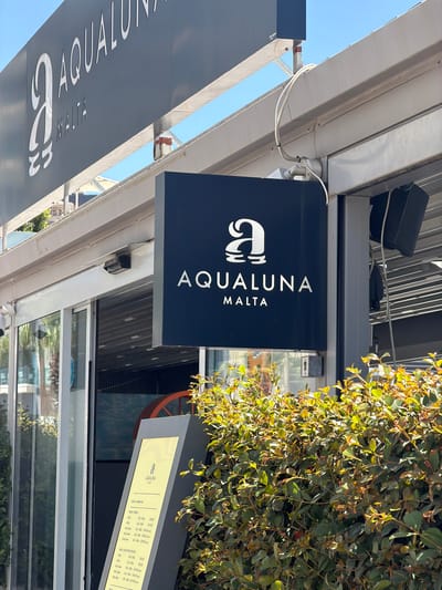 The image shows the exterior of a building with signage for "AQUALUNA MALTA". The building appears to be a commercial establishment, possibly a restaurant or a resort, given the name and the presence of outdoor seating and landscaping. One prominent sign is a dark blue rectangular sign with the "AQUALUNA MALTA" logo and text in white. Another, larger sign, likely the building's main facade signage, is visible above, also displaying "AQUALUNA MALTA" in white lettering on a dark grey background. The building has large glass windows and doors, suggesting an entrance. There is a display stand with a menu or information visible on the right side, partially obscured by lush green foliage. The scene is illuminated by natural daylight. The location is identified as Gżira, Malta.