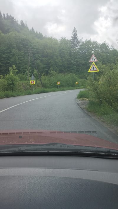 The image, likely taken from inside a vehicle, shows a road in a mountainous, forested area, possibly near Yasinya, Ukraine, as indicated by the road signs. The road surface is wet, suggesting recent or ongoing rain. Multiple road signs are visible along the side of the road, including a warning sign for "Dangerous curves" and several Chevron alignment markers. The weather is overcast and foggy. The interior of the vehicle is partially visible on the right side of the image, including parts of the dashboard.
