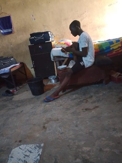 The scene depicts an interior space in Rumuekini, Nigeria. A male individual with dark skin and short hair is seated in profile, facing right, on a brown upholstered surface. He is wearing a white short-sleeved mesh shirt and black shorts with a white "DG" logo on the left thigh. His right foot is bare, while his left foot wears a blue flip-flop. He is looking down and manipulating a small, light-colored rectangular object with both hands. To his left, a small white plate with remnants is visible on a wooden structure.
The room features textured, light brown walls, exhibiting wear and discoloration. The words "SPACE B History" are handwritten in white on the upper right wall. A blue towel hangs from a hook on the upper left wall. The floor is concrete, displaying an uneven texture, dirt, and discolored patches. A lighter-colored, roughly rectangular patch is visible on the lower left floor.
Objects in the environment include:
- A black cabinet or wardrobe with silver handles, topped by a black hardshell suitcase with silver accents, located behind the seated individual.
- To the far left, another wooden stand holds a black soft-sided bag.
- On the floor, a black bucket with a silver handle is positioned left of the individual.
- A small silver pot with a lid rests near the bucket.
- A pair of dark-colored slip-on shoes are near the lower left corner of the frame.
- A multicolored patterned sheet or blanket (with yellow, blue, and red squares) and a white pillow are visible behind the seated individual.
- A red and white cardboard box is partially visible on the far right.
The room is illuminated by what appears to be a light source from the upper right, casting a bright highlight on the wall.