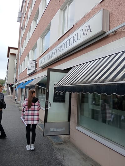 The image shows the exterior of a building with a business called "SALON LADY" and another sign that reads "VALOKUVAAMO SPOTTIKUVA". Two people are visible on the sidewalk in front of the building. One person, viewed from the back, is wearing a pink and white plaid jacket, black leggings, and grey sneakers, and is holding a rolled-up white paper. The other person, partially visible on the left, is wearing a dark jacket and appears to be walking.

The building has a pinkish-brown textured facade with multiple windows. A striped awning, black and white, is above the entrance to "VALOKUVAAMO SPOTTIKUVA". A sign on the glass door indicates operating hours from 10-16. Below this, there is smaller text, difficult to read, but it appears to include a website and a phone number. Blue awnings are visible further down the storefront. The sky is overcast, suggesting a cloudy day. The scene appears to be a commercial area in Kuopio, Finland.