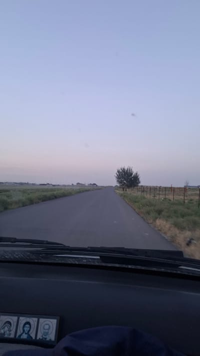 The image depicts a view from inside a vehicle, looking out onto a rural road in Chubarsu, Kazakhstan. The scene is captured at dusk or dawn, with the sky transitioning through pale blues and pinks. A paved road stretches into the distance, flanked by grassy fields and a fence line. A lone tree stands prominently on the left side of the road. The dashboard of the vehicle is visible in the foreground on the right, with a dark interior. On the dashboard, a small framed item with three portraits is visible, featuring stylized depictions of individuals. The overall impression is one of a quiet, perhaps solitary journey through a peaceful landscape. The time of day suggests either the beginning or end of daylight hours, creating a soft, diffused light across the scene. No specific activities or emotions are explicitly conveyed, beyond the implied movement of travel.