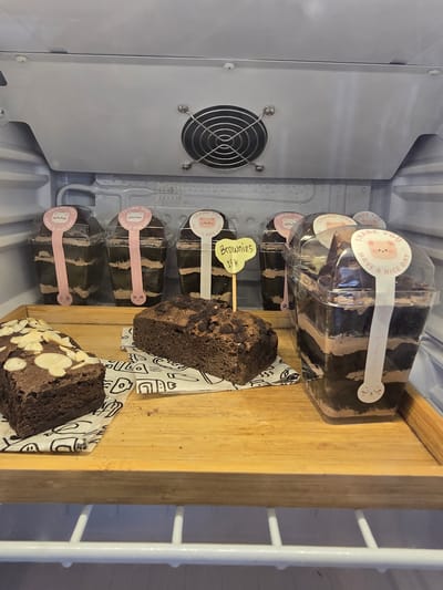 The image displays a close-up view of a refrigerated display case containing several chocolate brownies. On a wooden tray in the foreground, there are two pieces of brownie: one topped with sliced almonds and another with chocolate chips. Behind these, in clear plastic cups, are layered chocolate brownies with frosting. These cups are adorned with circular pink stickers featuring a cartoon bear and text that reads "THANK YOU HAVE A NICE DAY." One cup also has a yellow heart-shaped tag that says "Brownies 15K." The background shows the interior of a refrigerator, with a cooling fan visible at the top. The setting appears to be a commercial establishment, possibly a bakery or cafe, located in Klapanunggal, Indonesia. There are no people or any direct interactions visible in the scene.