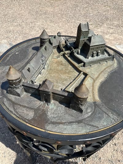 The image shows a detailed bronze model of Trakai Island Castle, situated on a circular platform. The model depicts the castle's walls, towers, courtyards, and prominent keep with its steep roof. The platform is supported by an ornate black metal stand with decorative scrollwork. The model is placed outdoors on a gravelly surface, likely in the vicinity of the actual castle in Trakai, Lithuania. The lighting suggests it is a sunny day, casting shadows across the model. There are no people visible in the image.