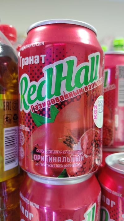 The image shows several red cans of a carbonated drink called "Real Hall." The cans are stacked and slightly tilted, with the prominent branding featuring green text and images of pomegranates. One can has a sticker with "KGZ" and a barcode, suggesting its origin or distribution in Kyrgyzstan. Text on the cans is in Cyrillic script and appears to be in Russian and possibly another language. One can shows the text "Гранат" (Granat), meaning pomegranate, and "Газированный напиток" (Gazirovannyy napitok), meaning carbonated drink. Another section of text reads "САЛҚЫНДАТЫЛҒАН КҮЙДЕЙШІҢІЗ ОРИГИНАЛЬНЫЙ" which translates to "Drink chilled, Original." The background is blurred but appears to be an indoor setting, possibly a store shelf or a collection of beverages. The lighting is bright, suggesting artificial light or a well-lit environment. The overall impression is a close-up product shot of a beverage brand. The presence of the Kyrgyzstan country code (KGZ) and the setting context suggest the location is Terek-Say, Kyrgyzstan.