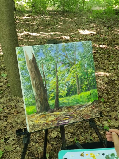 An oil painting on canvas, depicting a sun-dappled forest path with trees and foliage, is set up on a black tripod easel outdoors amidst a forest floor covered with fallen leaves. A person's hand, holding a paintbrush, is visible in the lower right corner, reaching towards the painting. A palette with various colors of paint sits below the easel. The lighting suggests it is daytime, with sunlight filtering through the trees. The scene appears to be in a wooded area.