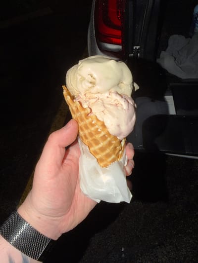 A close-up, low-angle shot shows a hand holding an ice cream cone with two scoops of ice cream. The ice cream is a light beige color and appears to have pieces of strawberry or raspberry mixed in. The cone is a golden brown waffle cone, wrapped in white paper. The hand is holding the cone from the bottom, with fingers wrapped around the paper. The background is dark, with a blurry image of a vehicle's taillight and the open trunk of a car. The ground appears to be asphalt. The scene is set in the city of Arlington Heights, United States of America.