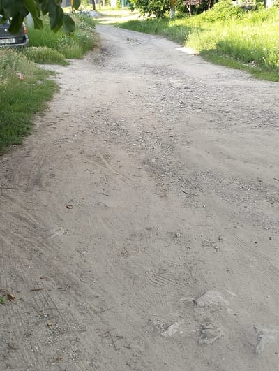 The image depicts a rural dirt road in Șoldănești, Moldova, likely during the daytime. The road surface is uneven, composed of gravel and dirt, with visible tire tracks and some scattered stones. Lush green grass and vegetation line the sides of the road, with trees and foliage in the background, suggesting a warm season. The light suggests it is either morning or late afternoon, with some shadows visible.

On the left side of the road, partially visible, is the rear of a dark-colored car, possibly a Renault Mégane, indicated by the visible badge. The license plate of the car reads "KTU 715". A plastic wrapper or bag is also visible in the grass near the car. The overall scene evokes a quiet, everyday rural setting. There are no people visible in the image, and no specific activity is taking place, other than the implied passage of vehicles on the road. The environment appears natural and somewhat unkempt, typical of a country lane.