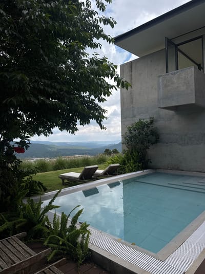 This image depicts a serene outdoor scene with a modern architectural element overlooking a lush landscape. The foreground features a swimming pool with clear, light blue water, reflecting the sky and surrounding greenery. The pool's edge is lined with a light-colored coping and a white grated drain. To the left of the pool, weathered wooden chairs are visible on a wooden deck.

Beyond the pool, a well-maintained green lawn is dotted with tall, wispy grasses. Two empty chaise lounges with light-colored cushions are positioned on the lawn, facing the distant view. In the mid-ground, a dense canopy of dark green leaves from a large tree overhangs the scene, with a single bright red hibiscus flower visible on its left side.

The background reveals a rolling landscape of hills covered in dense vegetation, fading into a hazy blue in the distance under a cloudy sky.

To the right of the pool, a striking modern structure made of concrete dominates the view. This building features clean lines, with a cantilevered section and large glass windows. A small green bush and ferns grow against the concrete wall near the pool.

While the image is labeled as being in Đa Lộc, Vietnam, there are no people visible in the scene, and no specific landmarks identifying it as being within a city. The overall impression is one of tranquility and luxurious seclusion, likely a private residence or resort.