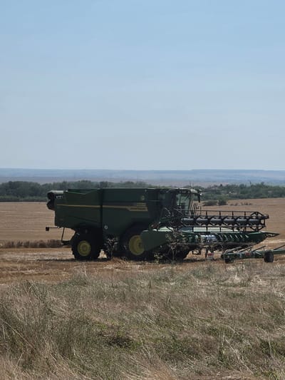 A John Deere combine harvester is parked in a dry, harvested wheat field under a clear, pale blue sky. The machine is green and yellow, with a large header attachment. The ground is covered in dry, brown grass and stubble. In the background, a line of trees can be seen, beyond which lies a hazy, undulating landscape with distant power lines. The scene appears to be in an rural area, likely on a sunny day. The image is a vertical shot, with the combine taking up a significant portion of the frame, angled slightly towards the viewer's right.