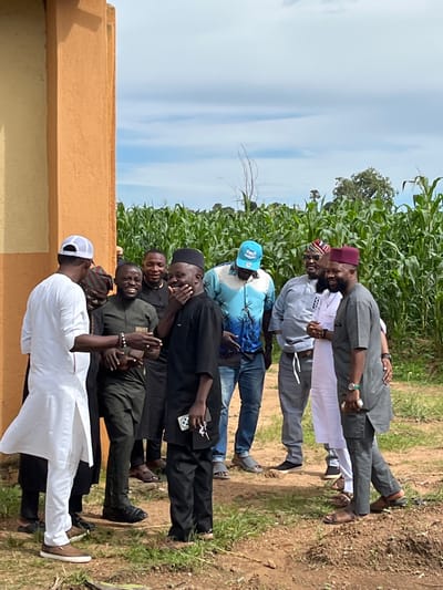 A group of seven Nigerian men are gathered outside near a building and a cornfield under a partly cloudy sky. The men are dressed in traditional clothing, with some wearing caps and others turbans. They appear to be engaged in conversation and social interaction, with some smiling and gesturing. The environment suggests a rural or semi-rural setting, possibly in Bawan Dodo, Nigeria, given the presence of the cornfield. The ground is dry and dusty, with some patches of green grass. The building on the left is painted a light orange color.