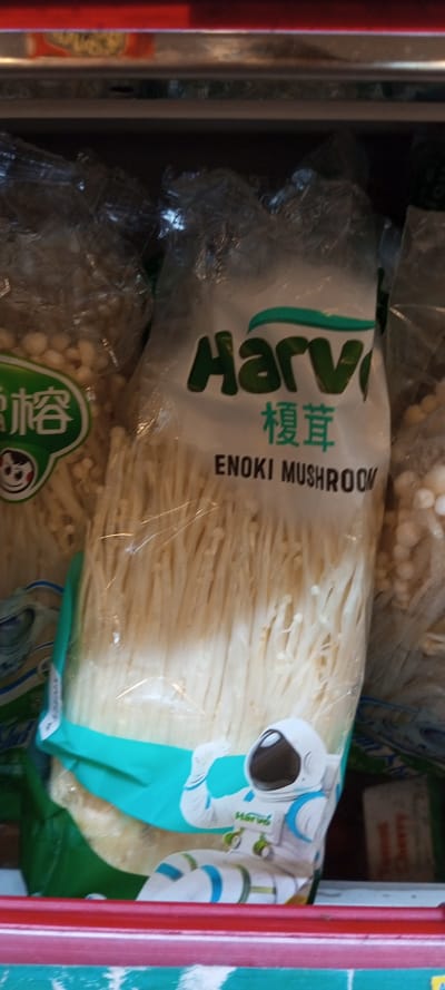 A package of "Harvi" brand enoki mushrooms sits on a shelf, next to other packages of similar mushrooms. The "Harvi" package features an astronaut illustration in the lower right corner. The mushrooms themselves are visible through the clear plastic packaging, showcasing their slender, white strands. The shelf below is red and green.
